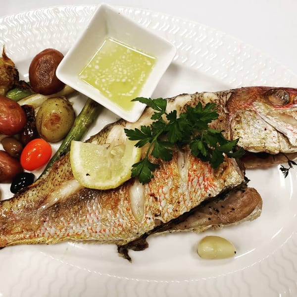 Whole roasted fish with lemon, parsley, roasted vegetables, and a side of sauce on a white plate.