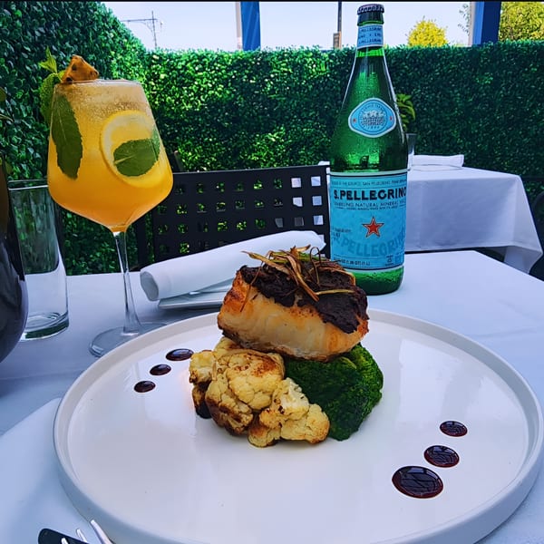 A fish dish with broccoli and cauliflower, a cocktail, and a bottle of S.Pellegrino on an outdoor table.