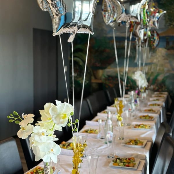 A long dining table set for a party with white orchids, balloons, and individual fruit salads.
