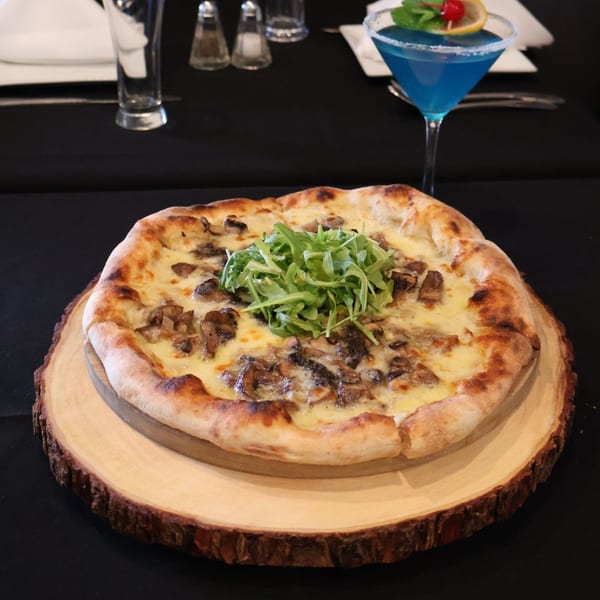 Mushroom pizza with arugula on a wooden board, with a blue cocktail in the background.