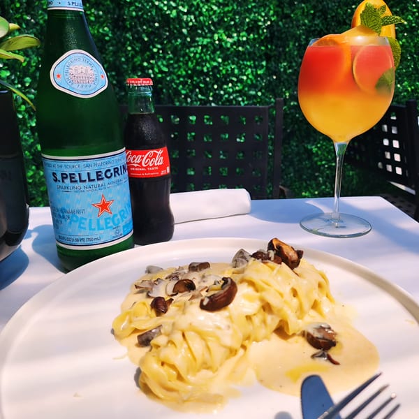 Creamy Italian pasta with mushrooms, S.Pellegrino, Coca-Cola, and a cocktail on an outdoor table.